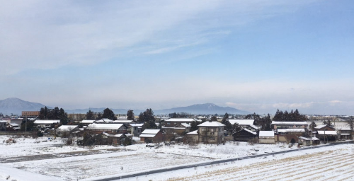 背景に山波が連なり、日本家屋の屋根には雪が積もっています。そして、日本家屋の前には雪にうっすら覆われた田んぼが広がっている景色の写真です。