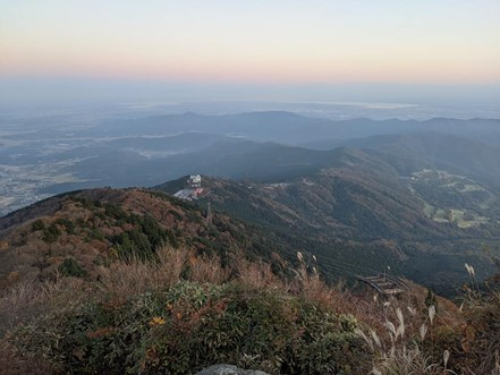 頂上から撮影されている。夕方早めの時間のうっすらとした夕やけと眼下には低い山脈が連なっている。すすきや紅葉した木々が山を彩っている。