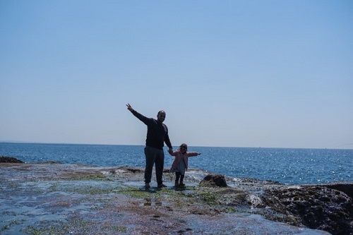 イフテカールさんとイナーヤちゃんが海岸の岩場で片手をあげてポーズしている