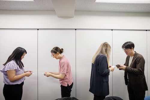 白い壁の前で、4人の人物が2つのペアに分かれて名刺交換をしている様子。