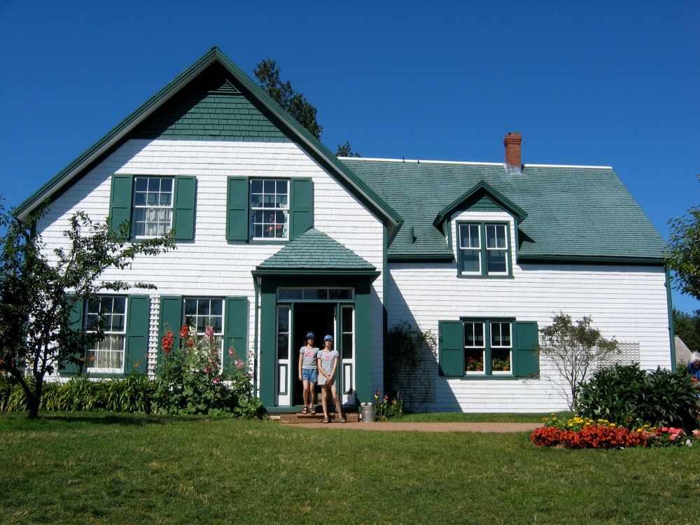 青空を背景にして、小さい頃のラターナス姉妹がプリンスエドワード島にあるアンの家（白いと緑色の家）の前に立っています。