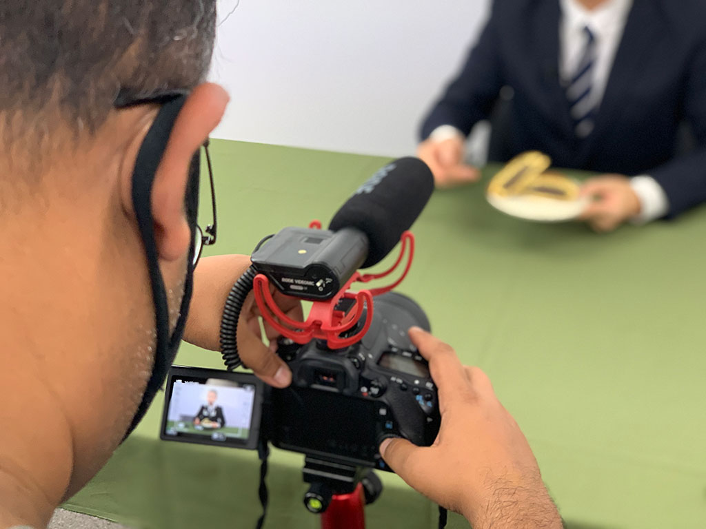 カメラマンのイフテカールがカメラのファインダーをみつめている様子で、奥にはホストのデキがどら焼きを持っている様子が映っています。